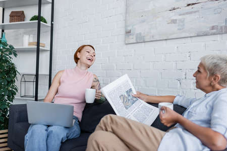 laughing woman sitting on couch with laptop near lesbian girlfriend with newspaper and cup of teaの写真素材