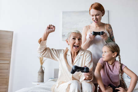 KYIV, UKRAINE - DECEMBER 8, 2021: Excited lesbian woman holding joystick near girlfriend and kid at homeの写真素材