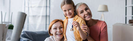 cheerful lesbian women looking at camera near adopted daughter holding key from new apartment, bannerの写真素材
