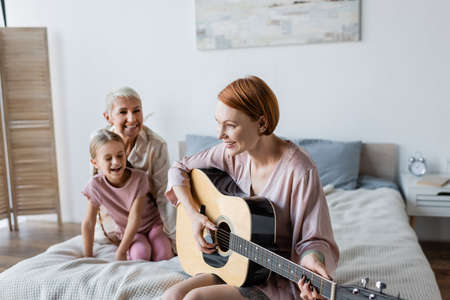 Happy woman playing acoustic guitar near blurred child and girlfriend on bed