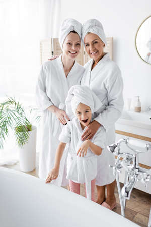 Positive lesbian couple in bathrobes hugging adopted daughter near bathtub in bathroomの写真素材
