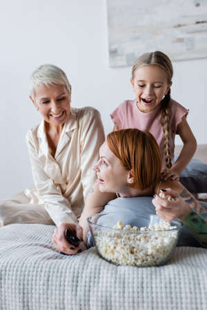Smiling lesbian woman holding popcorn and remote controller near kid and girlfriend on bed