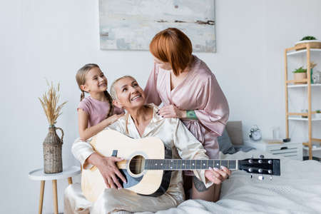 Cheerful lesbian woman playing acoustic guitar near child and girlfriend in bedroomの写真素材