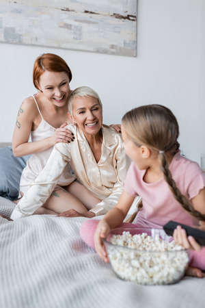 Happy lesbian couple hugging near kid with remote controller and popcorn on bed