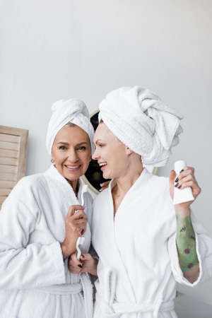 Positive lesbian couple in towels and bathrobes holding toothbrushes in bathroomの写真素材