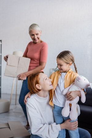 happy lesbian woman looking at adopted girl holding toy bunny in new apartment