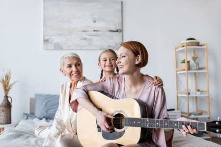 Smiling woman and kid looking at girlfriend playing acoustic guitar on bedの写真素材