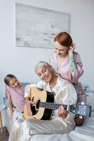 Smiling woman looking at girlfriend with acoustic guitar near daughter on bedの写真素材