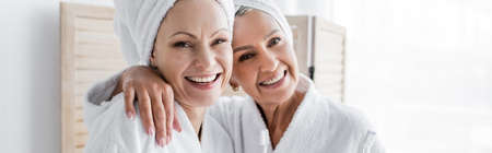 Smiling woman in bathrobe and towel hugging girlfriend with toothbrush at home, bannerの写真素材