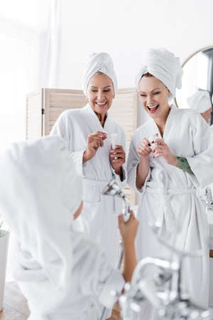 Positive lesbian couple holding creams near blurred adopted daughter with shower head in bathroomの写真素材