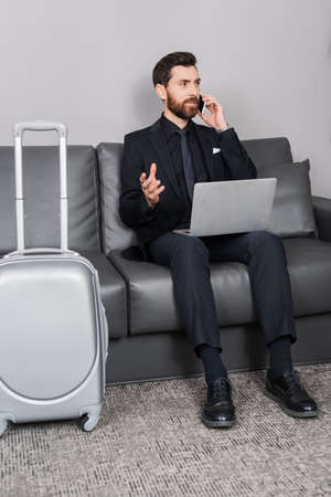 bearded businessman talking on smartphone near laptop and luggage in hotel roomの写真素材