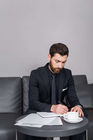 bearded man in suit writing on notebook near documents in hotel roomの写真素材