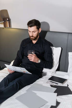 bearded man holding glass of whiskey and sitting in hotel bed near infographics