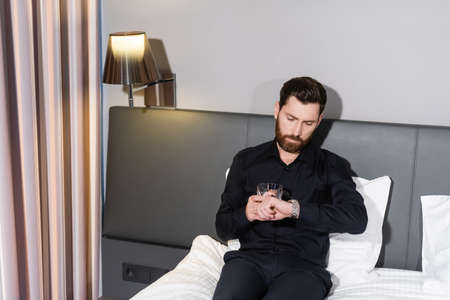 bearded man holding glass of brandy and checking time while sitting in hotel bedの写真素材
