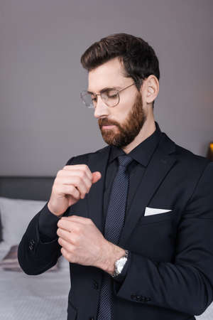 bearded man in eyeglasses adjusting sleeve of shirt in hotel room