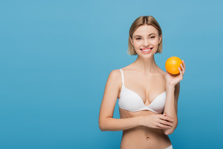 smiling young woman in white bra holding ripe orange isolated on blueの写真素材
