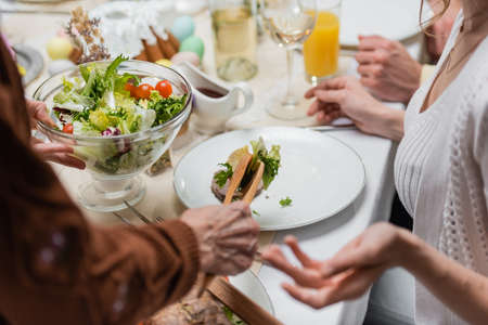 cropped view of woman holding bowl with fresh vegetable salad near blurred family having easter dinnerの写真素材
