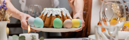 cropped view of woman holding plate with tasty easter cake and painted eggs, bannerの写真素材