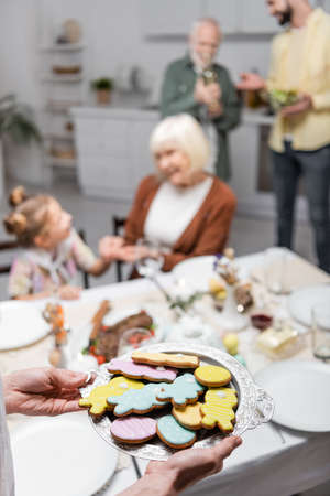 woman holding tray with traditional easter cookies near blurred family in kitchenの写真素材