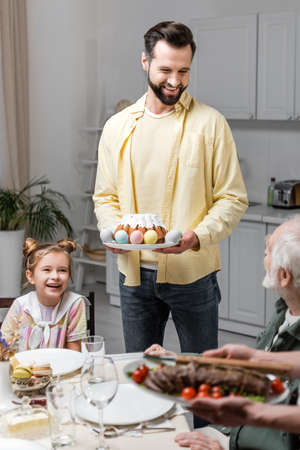 smiling man holding easter cake and painted eggs near family during festive dinnerの写真素材