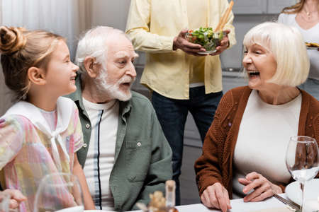 senior woman laughing near husband and granddaughter during family dinner