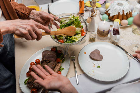 partial view of senior woman holding vegetable salad near meat on festive tableの写真素材
