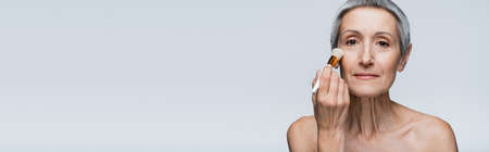 middle aged woman applying face powder with cosmetic brush isolated on grey, banner