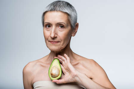 middle aged woman with bare shoulders holding ripe avocado isolated on greyの写真素材