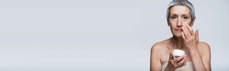 mature woman with grey hair holding container and applying cosmetic cream isolated on grey, bannerの写真素材