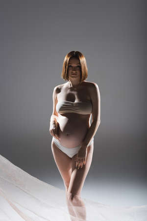 Pregnant woman in underwear posing near cloth on grey backgroundの写真素材