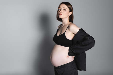 Stylish pregnant woman in top and jacket looking at camera on grey background