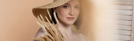 Woman in sun hat looking at camera near spikelets and folding screen isolated on beige, bannerの写真素材
