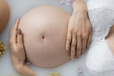 Cropped view of pregnant woman in bra touching belly in bath with flowers and milkの写真素材