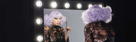 drag queen in purple wig holding cosmetic brush while looking in mirror, banner