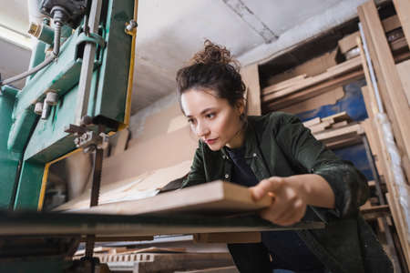 Brunette carpenter holding blurred plank near band saw in workshopの写真素材