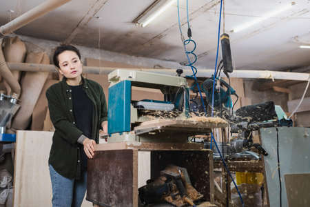 Young carpenter looking at blurred sawdust near thickness planer in workshopの写真素材