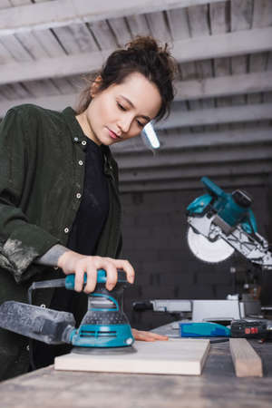 Carpenter using blurred sander on blurred plank near miter saw in workshop