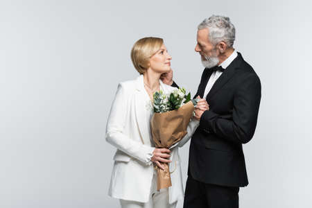 Side view of mature groom touching bride with wedding bouquet isolated on grayの写真素材