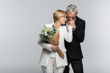 Mature groom kissing hand of bride in suit holding wedding bouquet isolated on grayの写真素材