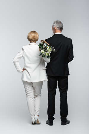Back view of bride in suit holding wedding bouquet near groom on gray backgroundの写真素材