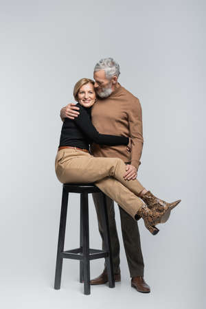 Cheerful stylish woman hugging mature husband while sitting on chair on gray backgroundの写真素材