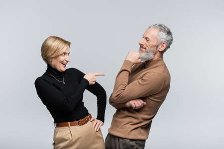 Positive mature woman pointing at stylish husband isolated on gray