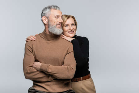 Smiling mature woman in turtleneck hugging husband and looking at camera isolated on grayの写真素材