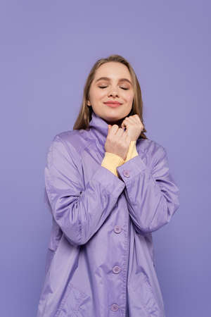 pleased young woman with closed eyes in trench coat isolated on purple