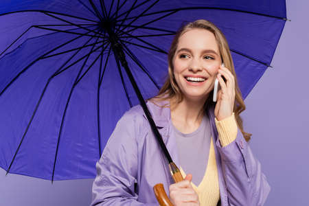 happy young woman in trench coat standing with blue umbrella and talking on smartphone isolated on...