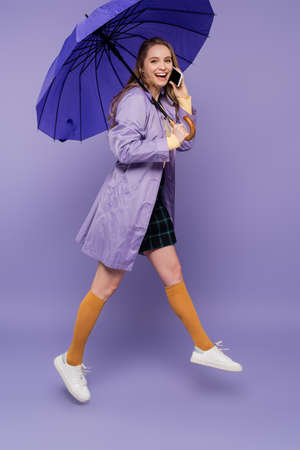 full length of happy young woman in trench coat walking with blue umbrella and talking on smartphone on purpleの写真素材
