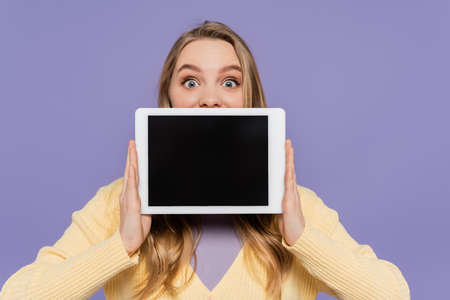 young woman covering mouth and holding digital tablet with blank screen isolated on purpleの写真素材