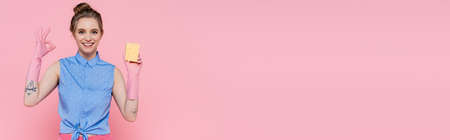 happy young woman holding sponge and showing ok sign isolated on pink, bannerの写真素材