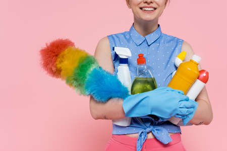 cropped view of pleased young woman in rubber gloves holding bottles with detergent and dust brush isolated on pinkの写真素材