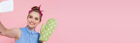 happy young woman with green baking glove taking selfie on smartphone isolated on pink, bannerの写真素材
